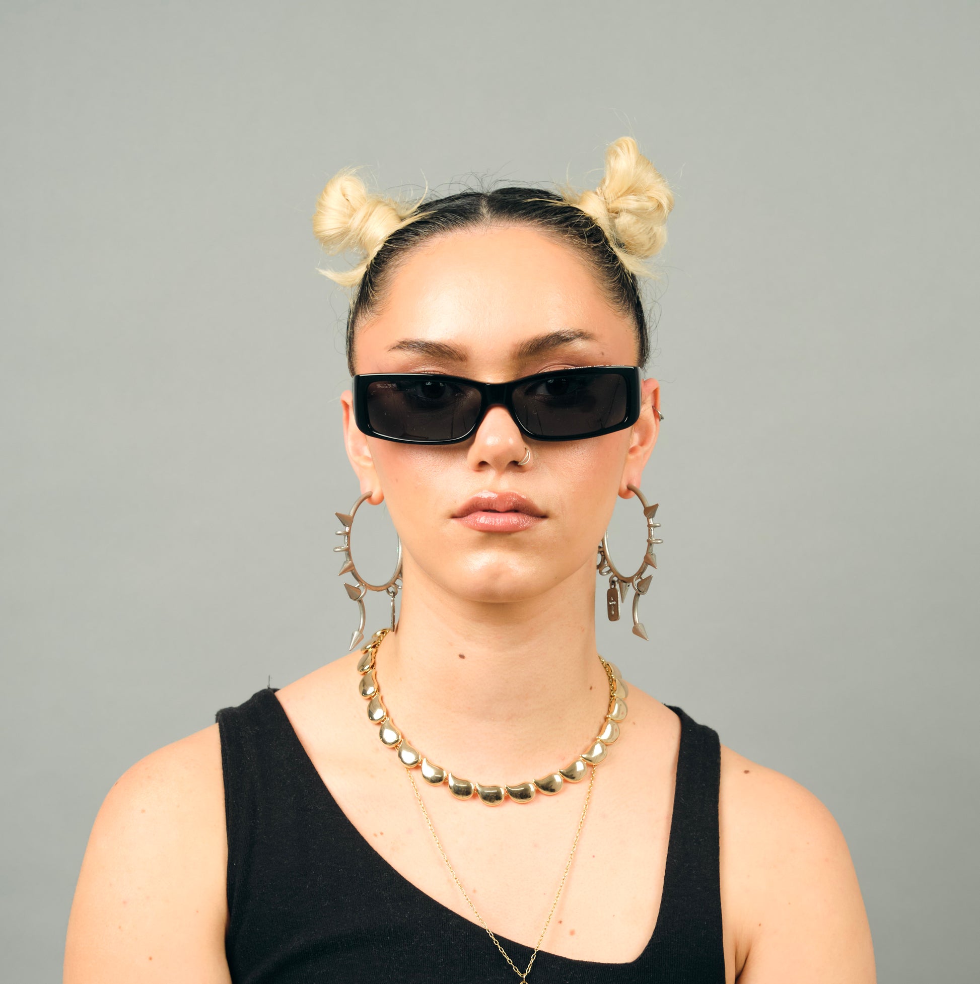Person wearing black sunglasses, a black top, and gold jewelry against a gray background